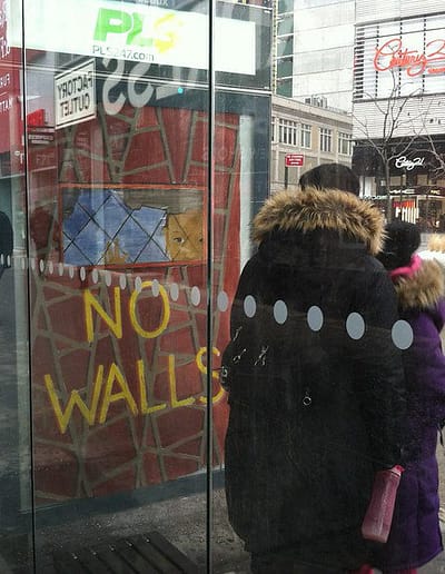 People wait in bus shelter on a city street where an ad box with the message, NO WALL is displayed.
