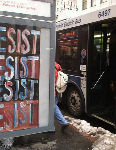 "Resist" appears four times in boldly painted letters and fills a poster inside an ad box at a city bus stop.
